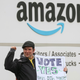 an amazon labour union organizer holding a sign that says "vote yes" stands in front of an amazon warehouse sign, with his fist raised